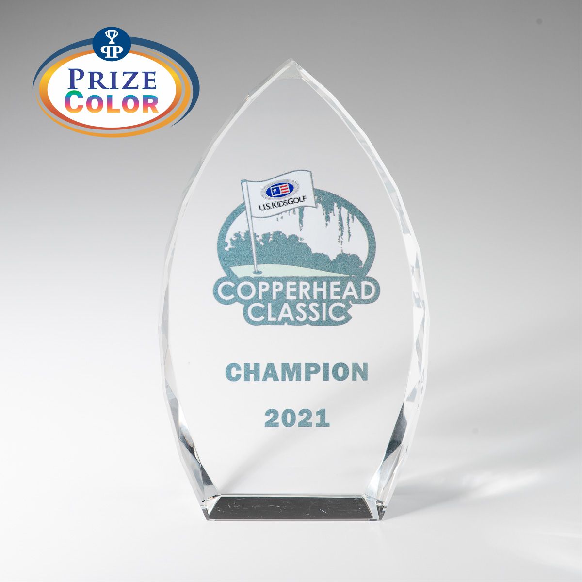 The Optic Crystal Award by Prize Possessions features clear glass with "Copperhead Classic Champion 2021" and a golf logo etched on it. A colorful Prize Color logo adorns the top left corner. Ideal for corporate awards or employee recognition. Plain white background.