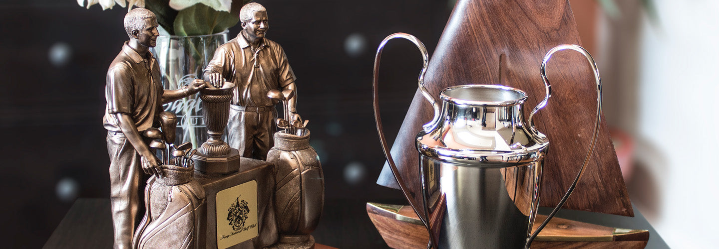 Several trophies are displayed on a table, including bronze statuettes of golfers with golf bags, a silver cup-shaped trophy with handles, and a wooden trophy shaped like a sailboat.