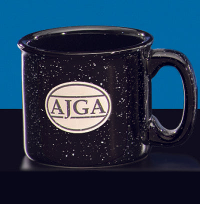 The Black Campfire Mug by Prize Possessions is a black speckled ceramic mug with a large handle and a white oval AJGA logo on the front, displayed against a blue and black background.