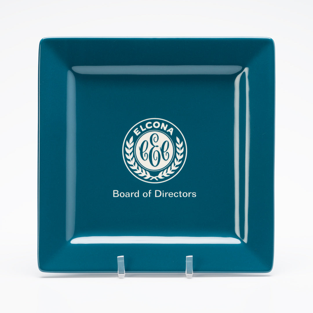 The Prize Possessions Blue Ceramic Square Plate features a white circular laurel and "ELCONA C&C" center, with "Board of Directors" below, in teal on a clear stand—ideal for modern tableware displays.