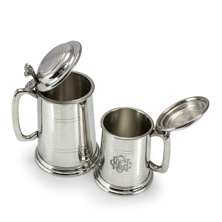 Two Prize Possessions British Pewter Lidded Tankards with large handles are side by side on a white background. One tankard features an engraved monogram; the other is plain. Both have shiny finishes and hinged lids.