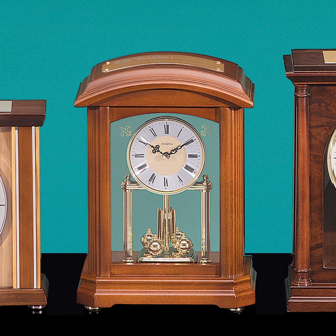 The Bulova Walnut Mantel Clock by Prize Possessions features a glass front, Roman numerals, gold accents, and a visible pendulum—an elegant choice for mantel or wood furniture décor, shown here flanked by two other wooden clocks.