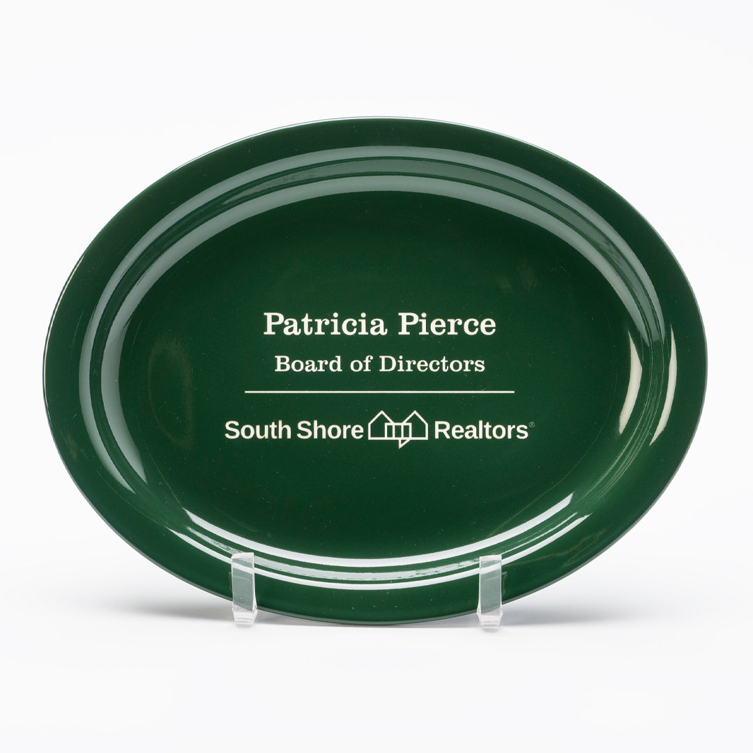 A Ceramic Green Wide Rim Platter by Prize Possessions, featuring "Patricia Pierce, Board of Directors, South Shore Realtors" and a house logo, sits on a clear stand as an elegant centerpiece against a white background.