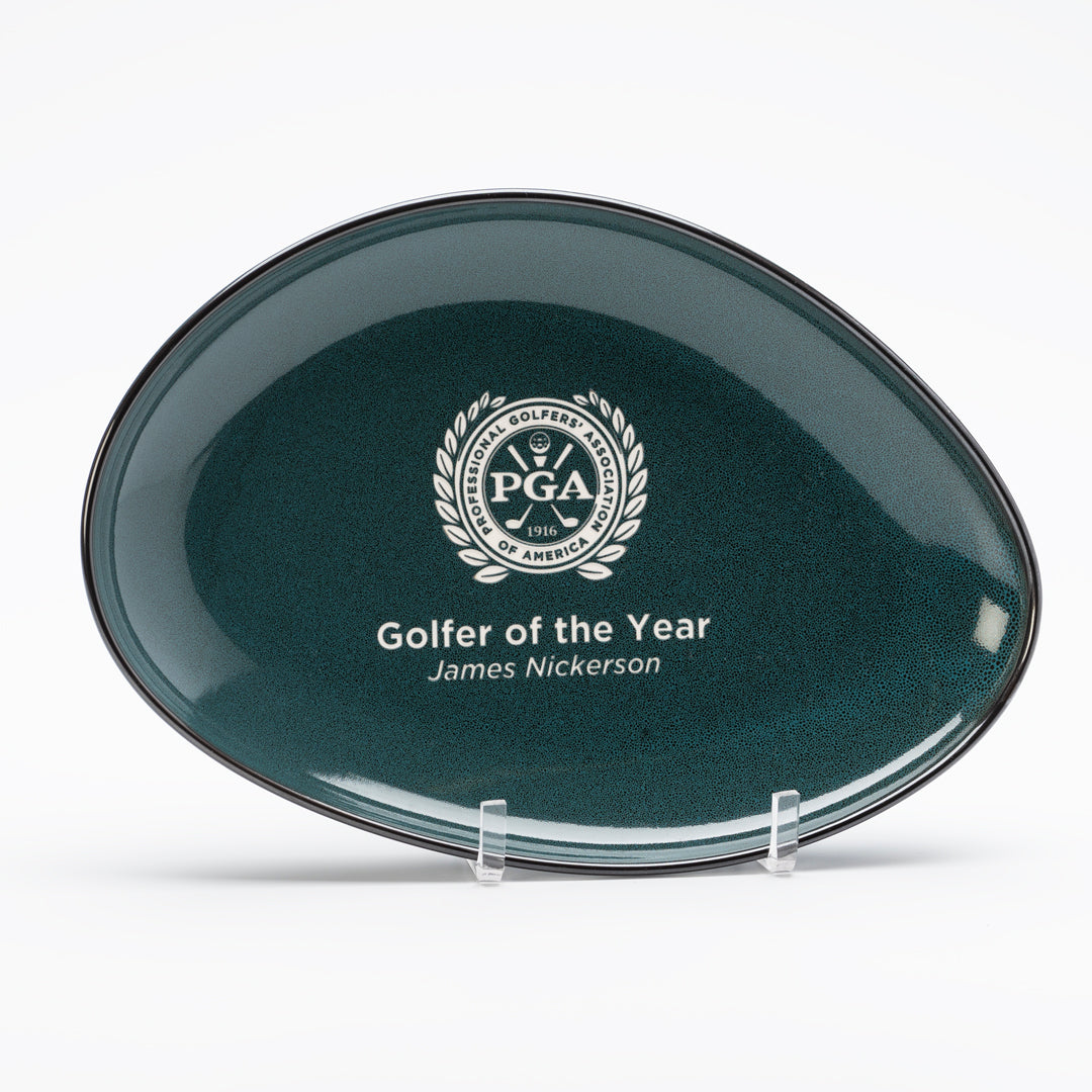 Ceramic Midnight Blue Luna Platter by Prize Possessions features the PGA logo and laurel wreath, with “Golfer of the Year, James Nickerson” engraved below. Resembling an elegant plaque, it is displayed on a clear stand.
