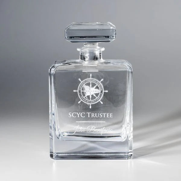 The Crystal Chateau Decanter by Prize Possessions is a clear whiskey decanter with a square stopper, engraved with a ship wheel design, "SCYC TRUSTEE," and John Bonifacio on the front.