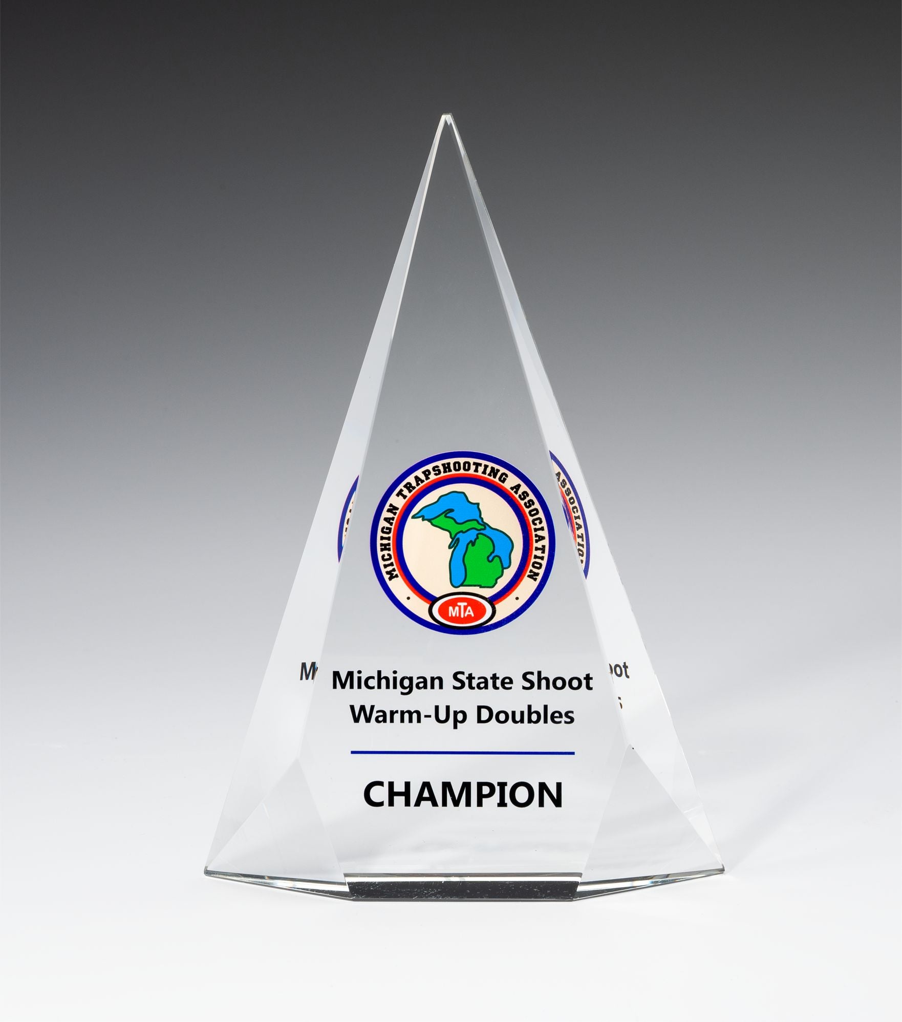 The Crystal Facet Triangle by Prize Possessions is a clear, pyramid-shaped award featuring the Michigan Trapshooting Association logo and "Michigan State Shoot Warm-Up Doubles CHAMPION" engraved on the front—ideal for recognition gifts.