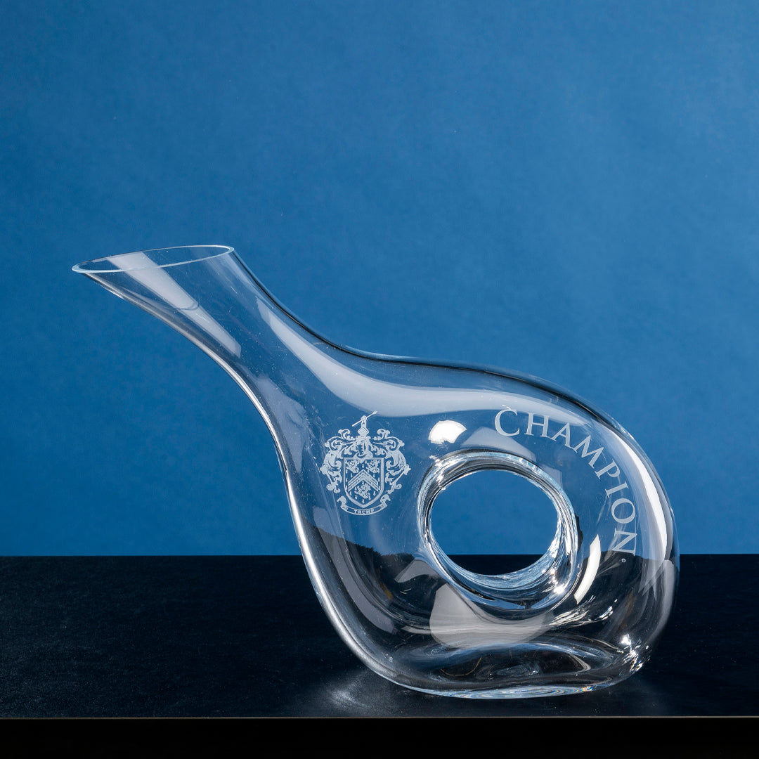 The Crystal Pierced Decanter by Prize Possessions, featuring a central round cut-out and engraved with a crest and "CHAMPION," rests on a dark surface against a blue background.