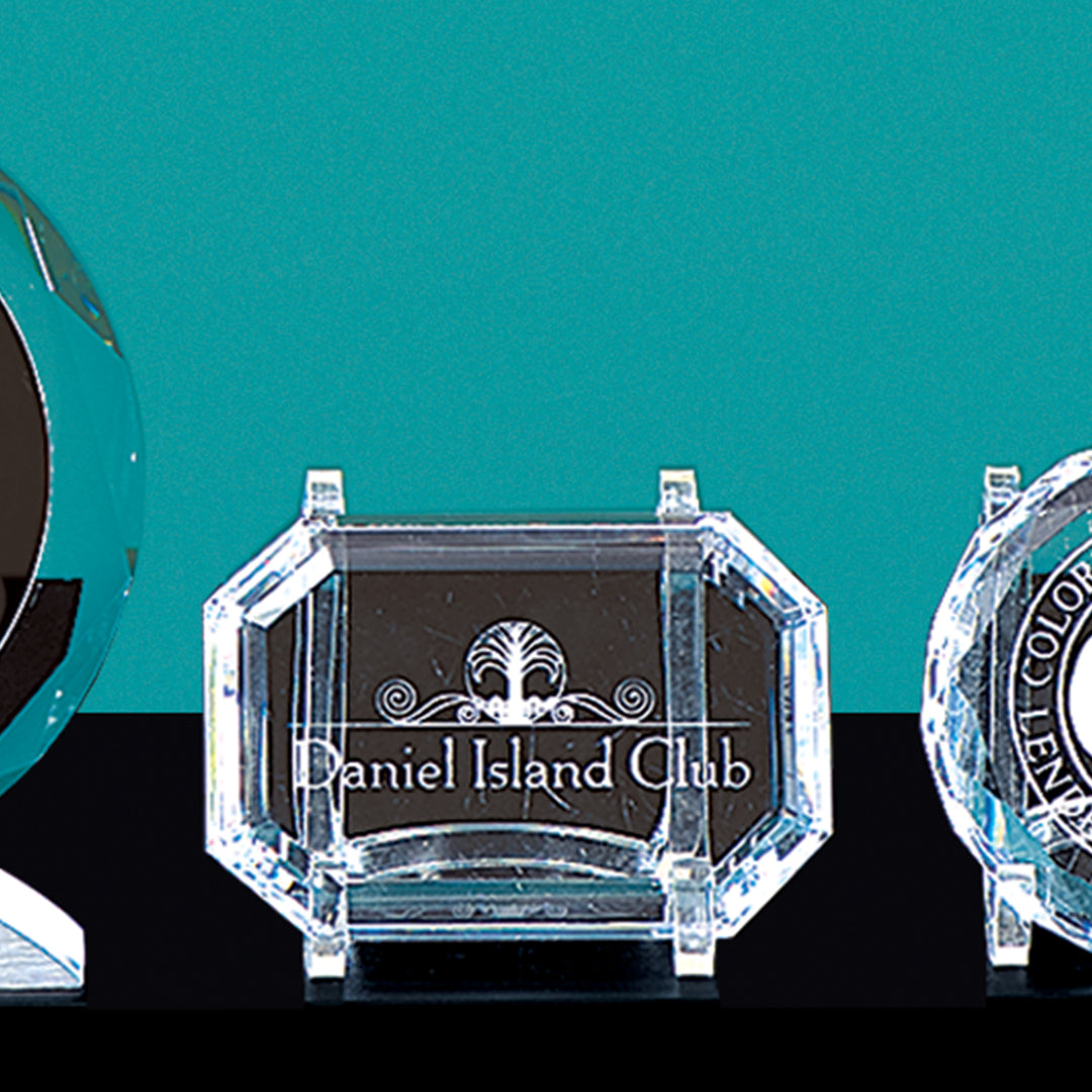 The Crystal Rectangular Paperweight by Prize Possessions is engraved with Daniel Island Club and a decorative emblem. Displayed against a teal background with other trophies, it makes an elegant office desk accessory or award.