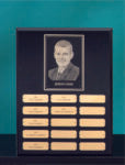 The Ebony Plaque with Zinc Profile by Prize Possessions displays a black-and-white engraved portrait at the top, gold nameplates in organized rows below, and a teal background for a refined, sophisticated look.