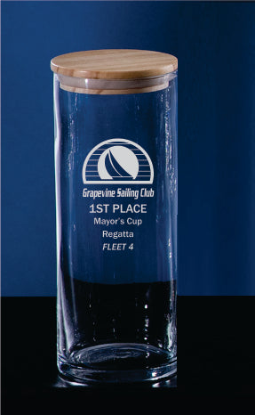 The Prize Possessions Glass Jar with Wood Cover, engraved with Grapevine Sailing Club 1st Place Mayors Cup Regatta Fleet 4 and a sailboat logo, is ideal for elegant kitchen storage and shown against a dark blue background.