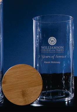 A clear Glass Jar with Wood Cover by Prize Possessions stands upright, featuring "Williamson College of the Trades, 5 Years of Service, Kevin Moloney" etched on it against a dark blue background.