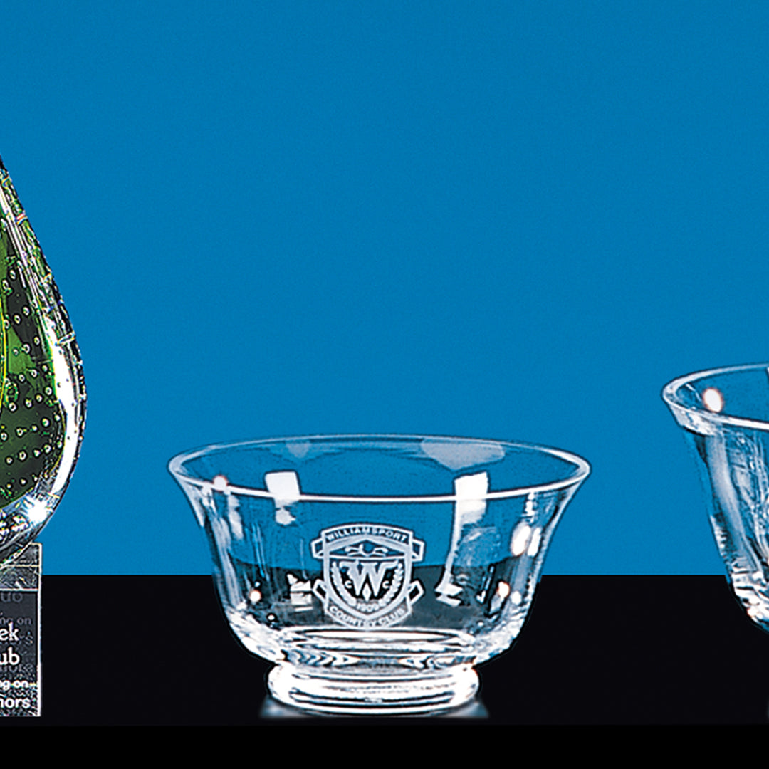 The Prize Possessions Glass Revere Bowl with an engraved emblem rests on a black surface against a blue backdrop, making a striking centerpiece, while parts of a green glass award and another bowl are partially visible nearby.