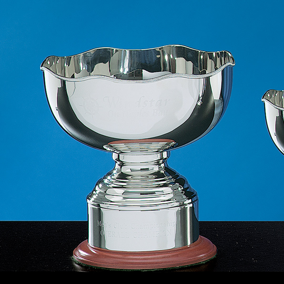 The Prize Possessions Nickle Plate Regency Bowl with Base features a shiny nickel finish and scalloped rim, set on a wooden base against a blue backdrop—ideal as an award or elegant centerpiece. Engraved text is partially visible on the cup.