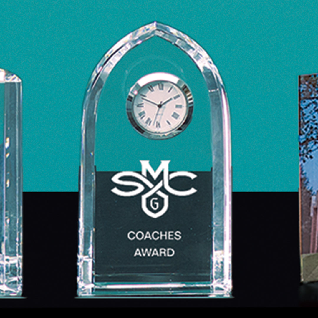 The Prize Possessions Optic Crystal Clock features a small round clock at the top with “SMC Coaches Award” engraved below, making it an elegant executive desk decor piece, displayed against a teal background.