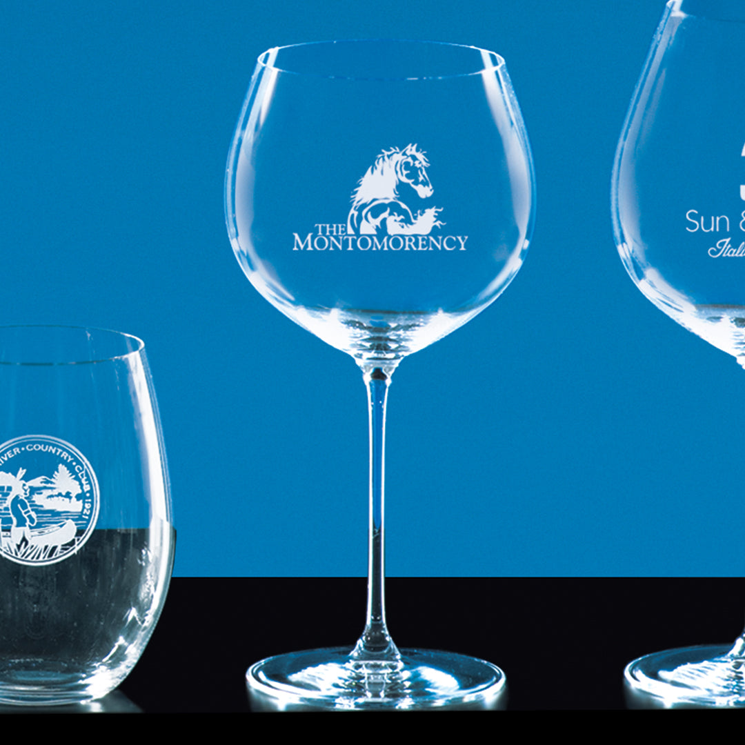 Three clear Riedel Oak Chardonnay glasses by Prize Possessions stand against a blue background; the center glass displays the Montomorency logo with a horse, while the other two also feature white emblems.