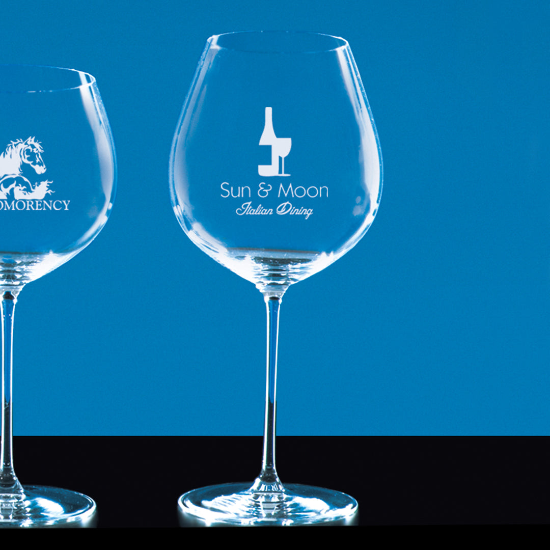 Two elegant crystal wine glasses by Prize Possessions: the right glass displays a Sun & Moon Italian Dining logo with a wine bottle, the left has part of a horse and "MORENCY"—perfect as Riedel Pinot Noir Glasses for wine tasting.