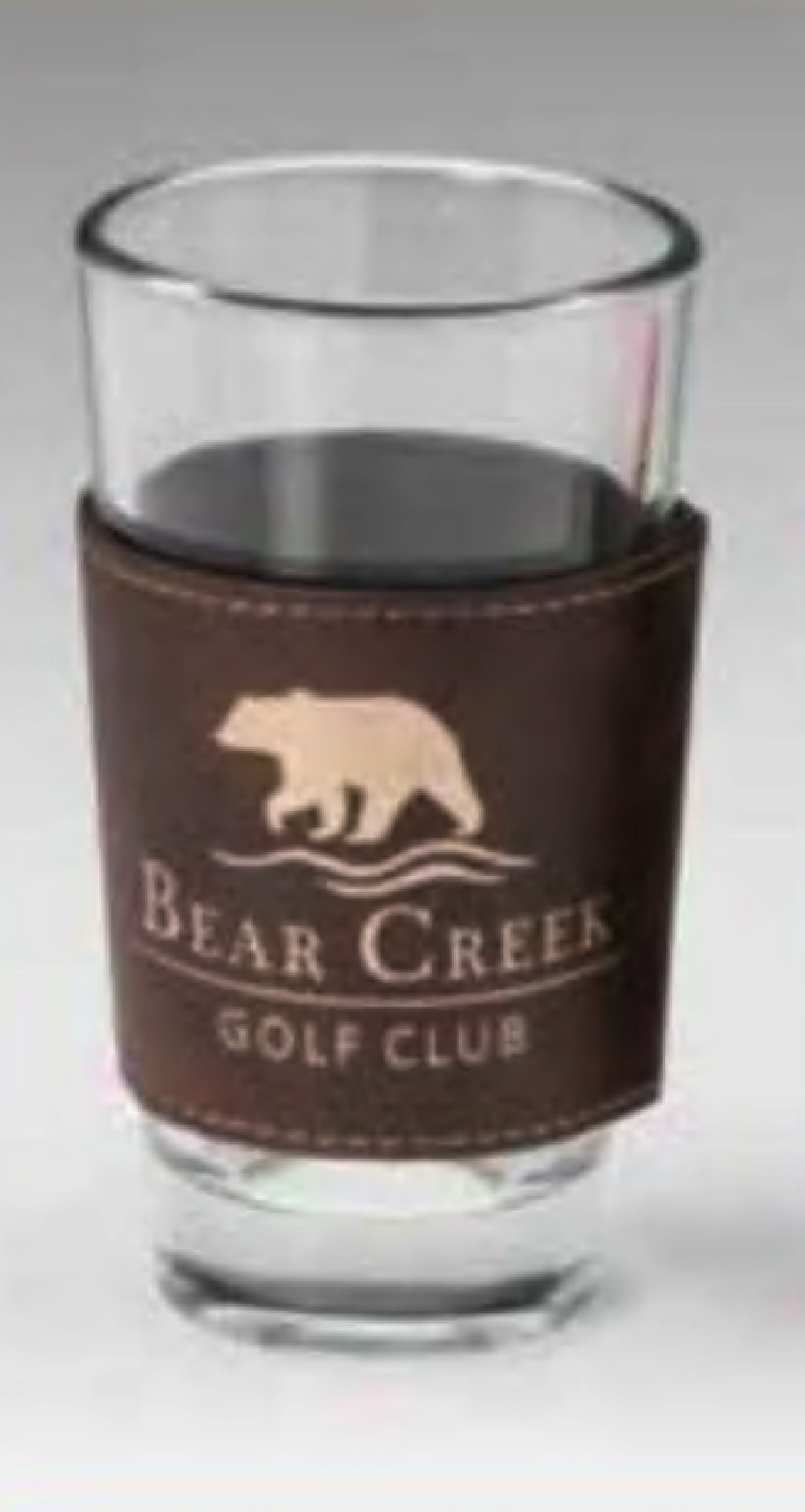 A clear glass with a dark drink is wrapped in the Rustic/Gold Leatherette Mug Sleeve by Prize Possessions, featuring "Bear Creek Golf Club" and an image of a walking bear above the text.