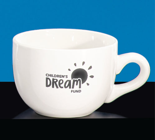 The Prize Possessions White Latte Mug with the Children’s Dream Fund sun logo sits on a black surface against a blue background—perfect for coffee lovers.