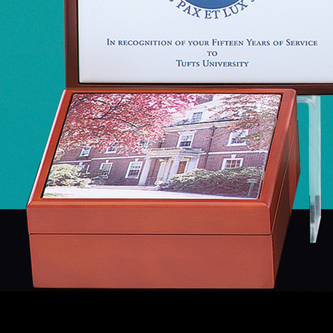 A Prize Possessions Wood Box with a ceramic tile lid featuring a photo of a building and flowering trees sits in front of a framed Tufts University fifteen-year service certificate.