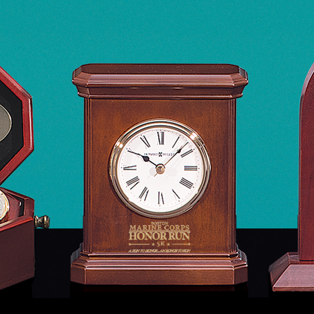 The Prize Possessions Wood Carriage Clock features a round white face with Roman numerals, gold rim, and an engraved "Marine Corps Honor Run 5K" plaque. Displayed on a black surface with a teal backdrop.