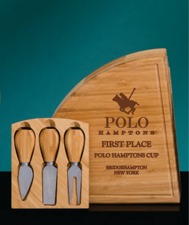 The Prize Possessions Wood Multi Level Cheese Board with Tools, featuring three cheese knives, a fork, and an engraved polo player logo with "POLO HAMPTONS FIRST PLACE POLO HAMPTONS CUP BRIDGEHAMPTON NEW YORK," is displayed on a green and black background.