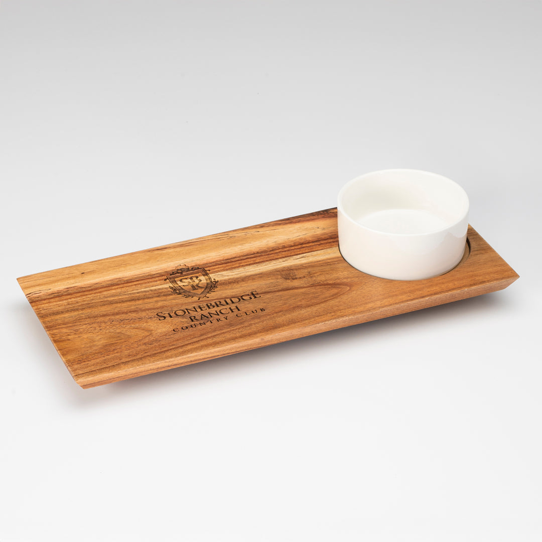 The Prize Possessions Wood Serving Board and Bowl Set features a rectangular board with an engraved logo, plus a recessed spot for the included round white ceramic bowl—ideal as elegant servingware or for charcuterie. Photographed on a white background.